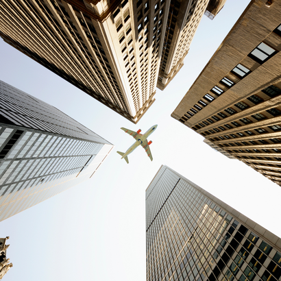 An airplane flying above tall office buildings in an urban setting, captured from the ground looking up at the sky.