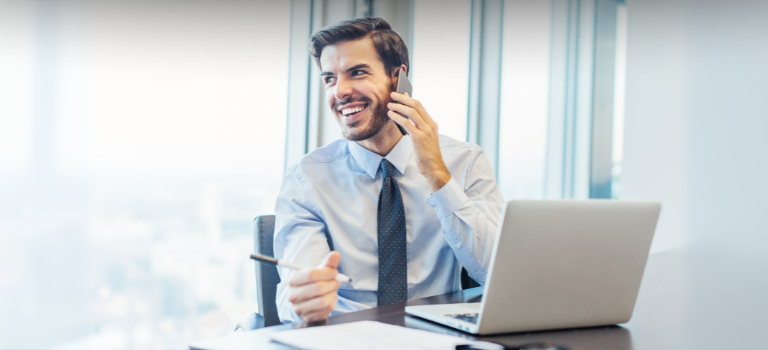 Мъж, който седи зад бюро с лаптоп пред него и телефон в ръка